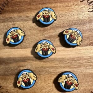 Six circular pins with a cartoon sailor flexing muscles. They are arranged on a wooden surface with engraved text around the edge.