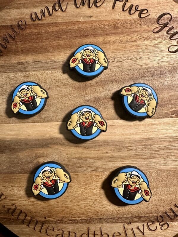 Six circular pins with a cartoon sailor flexing muscles. They are arranged on a wooden surface with engraved text around the edge.