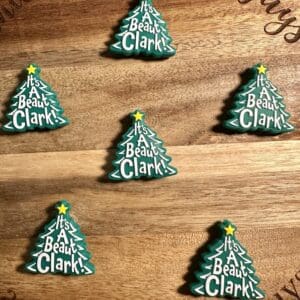 Seven Christmas tree-shaped cookies with the phrase "It's A Beaut Clark!" on a wooden surface.