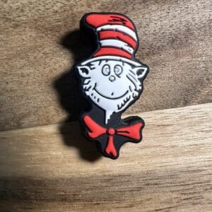 A lapel pin featuring a whimsical cat face with a red and white striped hat and a red bowtie, against a wooden background.