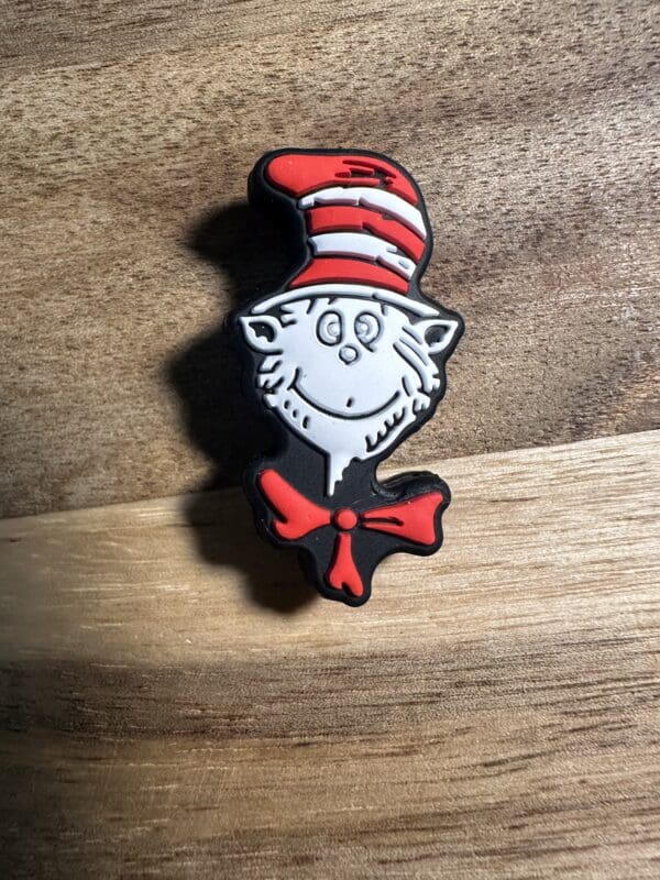 A lapel pin featuring a whimsical cat face with a red and white striped hat and a red bowtie, against a wooden background.