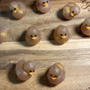 Ten small, glittery duck-shaped soaps arranged on a wooden surface.