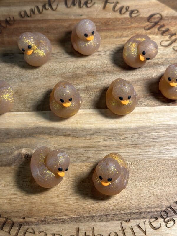 Ten small, glittery duck-shaped soaps arranged on a wooden surface.