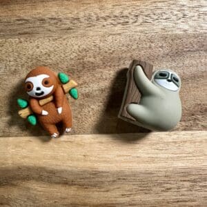 Two sloth-shaped erasers on a wooden surface, one is brown holding leaves, the other is gray climbing a log.