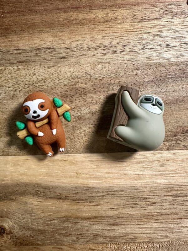 Two sloth-shaped erasers on a wooden surface, one is brown holding leaves, the other is gray climbing a log.