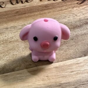 A small pink rubber toy shaped like a pig sits on a wooden surface with engraved text partially visible in the background.