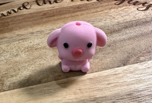 A small pink rubber toy shaped like a pig sits on a wooden surface with engraved text partially visible in the background.