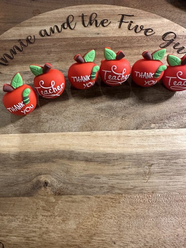 Five red apple-shaped ornaments with "Thank You" and "Teacher" written on them are displayed on a wooden surface with "Annie and the Five Guys" engraved above.